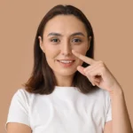 Young woman pointing to her nose while smiling against a neutral beige background.