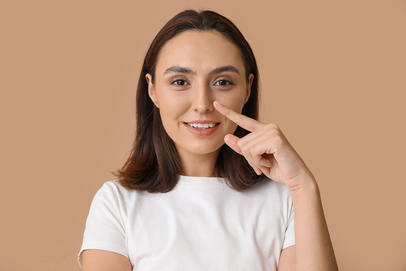 Young woman pointing to her nose while smiling against a neutral beige background.